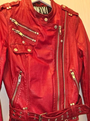 Röd skinnjacka från Nofama - Snygg röd skinnjacka från Jofama i bikerstil, flera dragkedjor och detaljer i silver. Jackan har bälte i midjan och snedställd dragkedja framtill. Perfekt för dig som vill sticka ut med en cool och look.