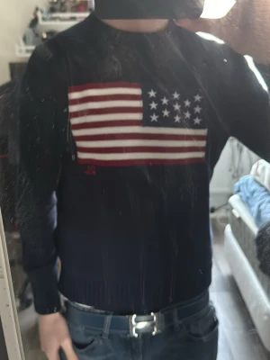 Mörkblå stickad tröja med USA-flagga - Säljer en mörkblå stickad tröja med en stor amerikansk flagga på bröstet. Tröjan har rund hals, långa ärmar och ribbstickade muddar. Perfekt för dig som vill ha en statement-piece i garderoben. Fler frågor finns här 😁