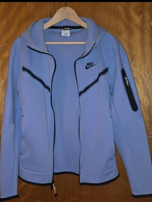 Nike tech hoodie med dragkedja - Nike tech hoodie i ljuslila med svart dragkedja och detaljer. Har huva, två sidofickor och en extra ficka med dragkedja på ärmen. Perfekt för en sportig och avslappnad stil. 