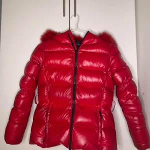  Colmar dunjacka med päls luva och bälte   - Snygg röd dunjacka från Colmar med fluffig huva och dragkedja framtill. Jackan har en quiltad design och en elastisk midja för bättre passform. Perfekt för kyliga dagar med stil.