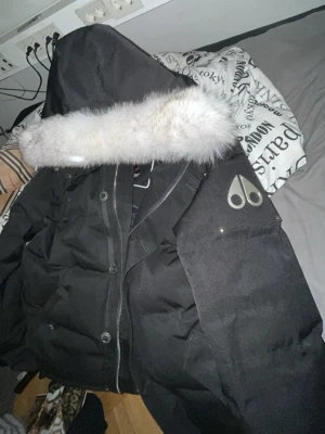 Svart dunjacka från Moose Knuckles - Säljer en svart dunjacka från Moose Knuckles med stor huva och vit fuskpäls. Jackan har en klassisk logga på ärmen, dragkedja och tryckknappar framtill samt flera fickor. Perfekt för kalla vinterdagar och riktigt varm.