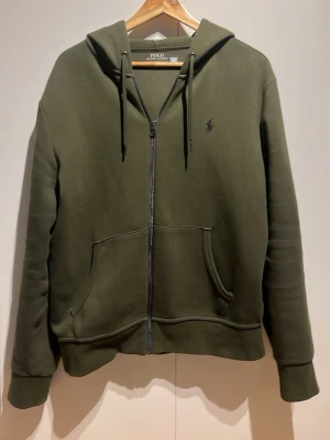 Olivgrön hoodie från Polo Ralph Lauren - Snygg olivgrön hoodie med dragkedja från Polo Ralph Lauren. Använd en gång, riktigt bra skick inga fläckar pris kan diskuteras. Kan skicka fler bilder om behövs!
