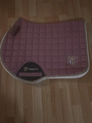 Rosa schabrak från Trinity - Säljer ett snyggt rosa schabrak från Trinity med quiltat mönster och glittrig kant. Märket syns tydligt på båda sidor och det finns kardborreband för enkel fastsättning. Perfekt till dig som vill ha lite extra stil i stallet.