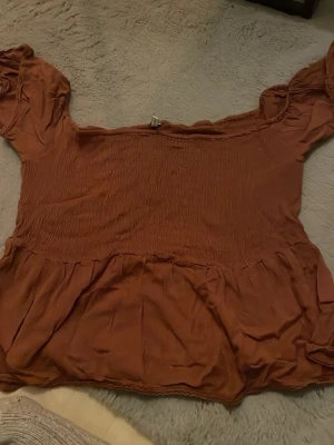 Brun offshoulder blus med volang - Säljer en brun blus med offshoulder-design och puffiga korta ärmar. Toppen har smock över bysten och en volang nedtill som ger en söt och trendig look. Perfekt att styla med jeans eller kjol för en chill vibe. Den är i strl 38 men är extremt stror på mig som vanligtvis har m/l så skulle mer säga att den passar xl/xxl 