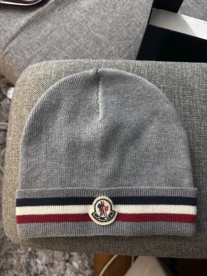 Grå Moncler mössa Tricolor - Snygg grå stickad mössa från Moncler med klassiska ränder i marinblått, vitt och rött vid kanten. Mössan har Moncler-loggan framtill och är tillverkad i mjukt material som sitter skönt på huvudet. Perfekt accessoar för kyliga dagar.