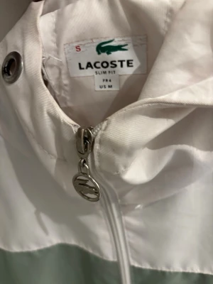 Lacoste vindjacka med huva - Snygg vindjacka från Lacoste i slim fit-modell. Jackan har färgblock i vitt, ljusgrönt och svart samt en praktisk huva. Dragkedja framtill med Lacoste-logga på zip-pullern. Perfekt för dig som vill ha en stilren och sportig look.