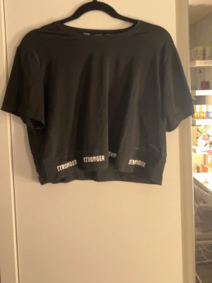 Svart croppad t-shirt från Stronger - Svart croppad t-shirt från Stronger med korta ärmar och rund halsringning. Nedtill finns vit text med loggan 'STRONGER' tryckt flera gånger. Perfekt för en sportig och avslappnad stil.