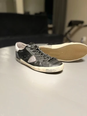 Svarta sneakers från Philippe Model - Snygga svarta sneakers från Philippe Model med vita detaljer och grå patch på sidan. Skorna har klassisk snörning och är tillverkade i skinn med en vit sula. Perfekta för dig som gillar en stilren och trendig look.