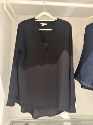 Svart v-ringad blus  - Stilren svart blus från Amisu med v-ringning och lång ärm. Blusen har en lös passform och är tillverkad i ett tunt, lätt material som ger ett snyggt fall. Perfekt att matcha med jeans eller kjol för en trendig look.