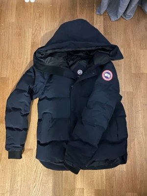 Mörkblå dunjacka från Canada Goose - Säljer en mörkblå dunjacka från Canada Goose med klassisk patch på ärmen. Jackan har stor huva, breda kanaler och ribbade muddar vid ärmslut. Perfekt för kalla vinterdagar och riktigt varm. Snygg och tidlös design som funkar till allt.
