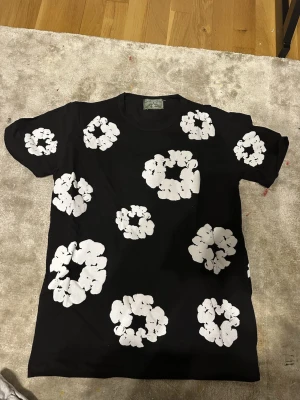 Svart t-shirt med vita blommor - Cool svart t-shirt från Stüssy med stora vita blommor tryckta över hela tröjan. Klassisk passform och rund halsringning, perfekt för en avslappnad streetwear-look.
