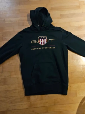 Mörkgrön hoodie från GANT - Mörkgrön hoodie från GANT med broderad logga och sköld på bröstet samt texten 'American Sportswear'. Klassisk känguruficka och justerbar huva. Perfekt för en avslappnad och sportig stil.