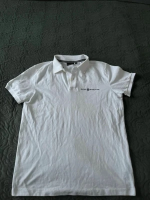 Vit pikétröja från Sail Racing - Klassisk vit pikétröja från Sail Racing med korta ärmar och krage. Tröjan har knappar framtill och en diskret logga på bröstet. Perfekt till en avslappnad och stilren look.