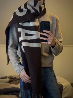Brun halsduk från Acne Studios - Säljer en stor och mjuk halsduk från Acne Studios i brunt med stora vita bokstäver. Halsduken har fransar i ändarna och är tillverkad i ett tjockt, värmande material som passar perfekt för kalla dagar. Snygg accessoar som sticker ut med sin grafiska design.