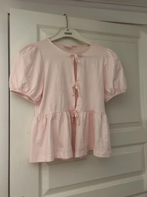 Ljusrosa blus med knyt från Gina Tricot - Supersöt ljusrosa blus från Gina Tricot med tre knyt framtill och volang nertill. Blusen har korta puffärmar och är lätt croppad, perfekt för en söt och trendig look. Tillverkad i mjukt material som känns skönt mot huden.