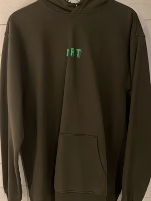 Mörkgrön hoodie med FRT-tryck - Snygg mörkgrön hoodie från FRT med liten grön logga på bröstet. Klassisk modell med huva och stor magficka framtill. Perfekt för dig som gillar en clean och stilren look. Mjuk insida och ribbade muddar vid ärmslut och nederkant.