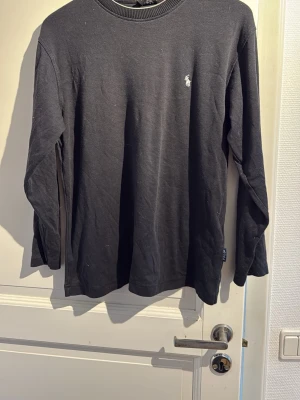 Svart långärmad tröja från Polo Ralph Lauren - Klassisk svart långärmad tröja från Polo Ralph Lauren med den ikoniska vita logobrodyr på bröstet. Tröjan har rund halsringning och är gjord i mjuk bomull som känns skön mot huden. Perfekt för en clean och stilren look.