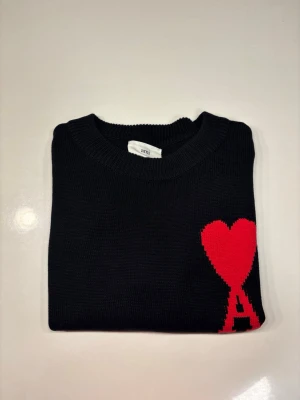 Svart stickad tröja med rött hjärta - Svart stickad tröja från AMI Paris med ett stort rött hjärta och bokstaven A på bröstet. Tröjan har rund halsringning och är tillverkad i ett mjukt, tjockt material som passar perfekt för kyligare dagar.