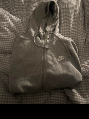 Grå zip-hoodie från Nike - Grå hoodie från Nike med dragkedja framtill och klassisk huva med dragsko. Tröjan har två stora fickor på magen och en vit Nike-logga på bröstet. Perfekt för en avslappnad och sportig stil. Nypris är runt 500! 