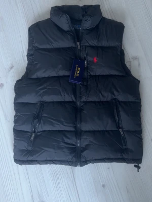 Svart dunväst från Polo Ralph Lauren - Stilren svart dunväst från Polo Ralph Lauren med röd broderad logga på bröstet. Västen har två dragkedjefickor framtill och hög krage. Perfekt för lager-på-lager och håller dig varm med sin vadderade design.