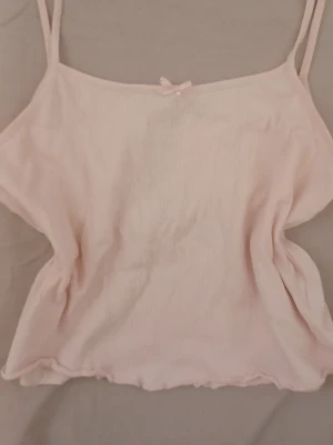 Ljusrosa linne med rosett - Sött ljusrosa linne med tunna axelband och en liten satinrosett framtill. Tyget är mjukt och har en lätt rynkad struktur som ger en avslappnad känsla. Perfekt för mysiga hemmakvällar eller som loungewear.