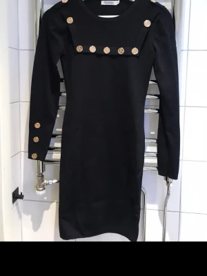 Svart klänning med guldknappar - Stilren svart klänning från Allusion med dekorativa guldknappar på axlar, bröst och ärmslut. Klänningen har lång ärm och en rak, kroppsnära passform som ger en elegant look. Perfekt för dig som gillar klassiska detaljer och enkel design.