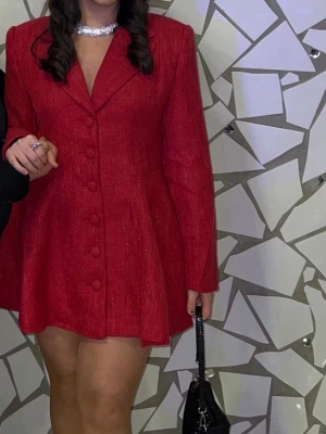Röd kavajklänning med knappar - Stilren röd kavajklänning med markerad midja och stora klädda knappar framtill. Klänningen har lång ärm och en lätt utsvängd kjol, perfekt för dig som vill sticka ut. Bär den med smycken och en liten väska för en trendig look. Passar perfekt till alla hjärtans dag. Funkar bra för korta och långa tjejer. Använts 1 gång, storlek 36/38.. Pris går att diskutera. 