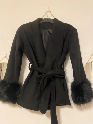 Svart pälsjacka med bälte - Stilren svart pälsjacka med fuskpälsdetaljer vid ärmsluten och knytbälte i midjan. Jackan har en klassisk krage och är helfodrad för extra värme. Perfekt för dig som vill ha en trendig och elegant look under vintern.
