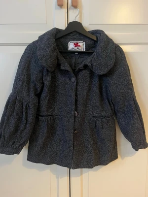 Grå ullkappa med stor krage - Snygg grå kappa från Noa Noa i ullblandning med stor, rundad krage och knappar framtill. Kappan har puffiga ärmar och markerad midja för en cool siluett. Perfekt för kyliga dagar när du vill vara både varm och stilren.