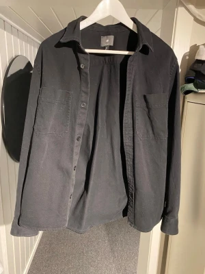 Svart skjorta från H&M med bröstfickor - Svart skjorta från H&M i regular fit med två bröstfickor och knäppning framtill. Skjortan har lång ärm och klassisk krage, perfekt för en stilren look. Tillverkad i mjukt bomullstyg som känns bekvämt hela dagen.