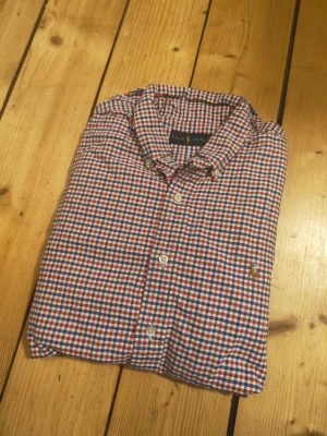 Rutig skjorta från Ralph Lauren - Klassisk rutig skjorta från Ralph Lauren i blått, rött och vitt. Skjortan har knappar framtill, lång ärm och en liten broderad logga på bröstet. Tillverkad i mjuk bomull och har en tidlös passform som funkar till många olika stilar.