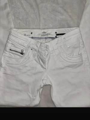 Vita jeans från River Island - Säljer ett par vita jeans från River Island med snygga detaljer som dragkedja på fickan och broderad logga på bakfickan. Klassisk femficksmodell med raka ben och silverfärgade nitar. 