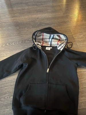 Svart hoodie med dragkedja och rutig huva - Svart hoodie från Burberry med dragkedja framtill och klassiskt rutigt foder i huvan. Tröjan har känguruficka och ribbade muddar vid ärmslut och nederkant. Perfekt för en stilren och avslappnad look.