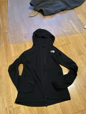 Svart vindjacka från The North Face - Svart vindjacka från The North Face med huva och hel dragkedja framtill. Jackan har två sidofickor med dragkedja och en diskret logga på bröstet. Tillverkad i ett lätt och smidigt material som skyddar mot vind. Perfekt för utomhusaktiviteter och streetstyle.