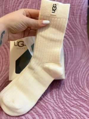 Ribbade vita strumpor från UGG - Säljer ett par ribbade vita strumpor från UGG. De är höga och har en mjuk och skön känsla, perfekta för att hålla fötterna varma. UGG-loggan sitter upptill på skaftet för en snygg detalj.