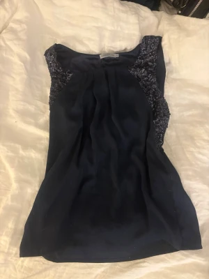 Mörkblå topp från Soaked in Luxury - Snygg mörkblå ärmlös topp från Soaked in Luxury med glittriga detaljer vid axlarna. Toppen har en lätt glansig finish och ett elegant fall. Perfekt att styla med jeans eller kjol för en trendig look.