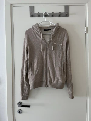 Beige ziphoodie från Lexington - Snygg beige hoodie från Lexington med dragkedja framtill och huva med dragsko. Mjuk bomullsblandning och broderad Lexington-logga på bröstet. Perfekt för en avslappnad och stilren look.