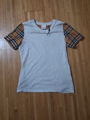 Vit t-shirt med rutiga ärmar från Burberry - Snygg vit t-shirt från Burberry med klassiska rutiga ärmar i beige, svart och rött. T-shirten har rund halsringning och är tillverkad i mjuk bomull. Perfekt för dig som vill ha en stilren look med en touch av lyx.