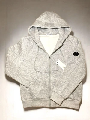 Ljusgrå hoodie från C.P. Company - Ljusgrå hoodie med dragkedja från C.P. Company. Klassisk modell med huva, känguruficka och ribbade muddar. Märkets ikoniska svarta linsdetalj sitter på ärmen. Tillverkad i mjuk bomullsblandning för extra komfort.