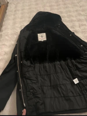 Svart parkasjacka från Bondelid - Snygg svart parkasjacka från Bondelid med mjukt foder och dragkedja framtill. Jackan har en klassisk design med huva och tryckknappar. Perfekt för kyliga dagar och riktigt skön att bära. Märkt med Bondelid-logga på insidan.