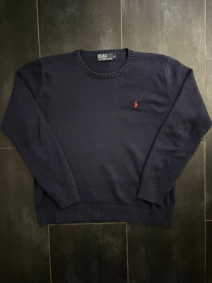 Mörkblå stickad tröja från Polo Ralph Lauren - Klassisk mörkblå stickad tröja från Polo Ralph Lauren med rund halsringning och ribbade muddar. Tröjan har den ikoniska röda broderade loggan på bröstet. Perfekt till jeans eller chinos för en clean look.