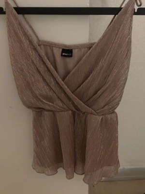 Glittrig omlottopp från Gina Tricot - Glittrig topp från Gina Tricot i ljusbrun nyans med omlott-design och smala axelband. Toppen har en lätt volang nertill och är perfekt för dig som vill sticka ut. Materialet är glansigt och ger en lyxig känsla.