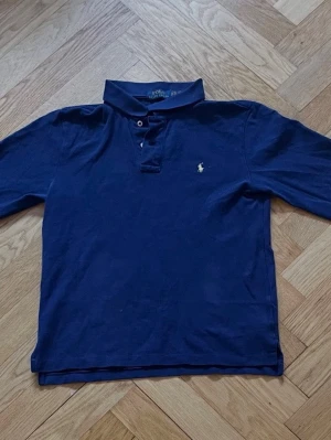 Ralph Lauren långärmade piké - Klassisk mörkblå henley från Polo Ralph Lauren med lång ärm och liten broderad logga på bröstet. Skjortan har knappar vid halsen och en mjuk bomullskänsla. Perfekt för en avslappnad men ändå stilren look.