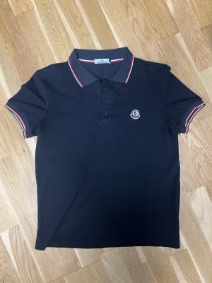 Svart Moncler pikétröja med röd/vita detaljer - Klassisk svart pikétröja från Moncler med röd och vit rand på krage och ärmslut. Tröjan har korta ärmar, knappslå framtill och Moncler-logga broderad på bröstet. Perfekt för en clean och sportig look.