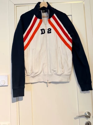 Vit och marinblå hoodie med D2-tryck - Snygg hoodie från Dsquared2 i vitt och marinblått med röda och vita ränder på bröstet. Dragkedja framtill, två fickor med dragkedja och hög krage. D2-tryck på bröstet ger en sportig vibe. Perfekt för dig som gillar streetwear och retrostil.