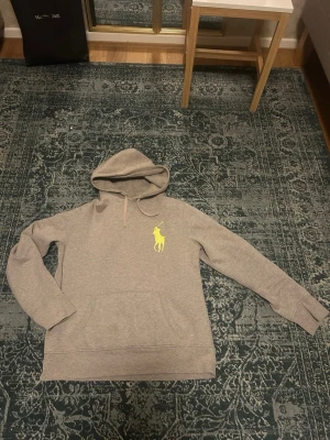 Beige hoodie från Polo Ralph Lauren - Snygg beige hoodie från Polo Ralph Lauren med stor gul logga på bröstet. Klassisk huva med dragsko och känguruficka framtill. Mjuk bomullsblandning som är perfekt för en avslappnad stil.