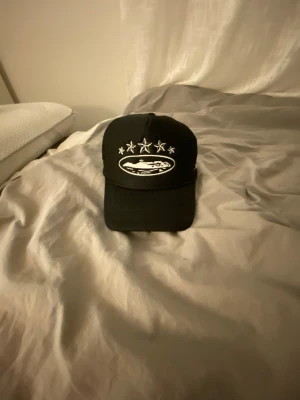 Svart truckerkeps med broderi - Svart truckerkeps med vit broderad design framtill som visar stjärnor och ett motiv av en bil. Kepsen har mesh på sidorna och bak för extra ventilation samt justerbar snapback. Perfekt för en streetwear-look.