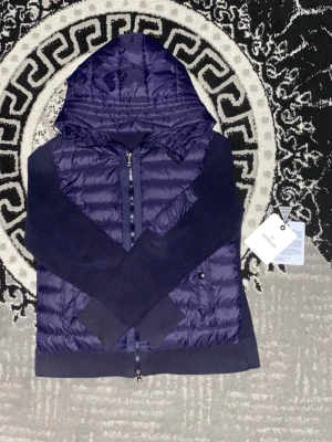 Mörkblå dunväst med huva från Moncler - Säljer en mörkblå dunväst från Moncler med stickade ärmar och stor vadderad huva. Västen har dragkedja framtill, ribbade muddar och en ficka med dragkedja på ärmen. Insidan har Moncler-etikett och serietidningsdetalj. Perfekt för dig som gillar exklusiva streetwear-plagg.passar båda killar och tjejer  Den e M men den passar S ochxå 