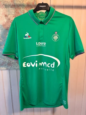 Saint Etienne LeCoqSportif 2016/17 kit, size L - Very good condition.  #saintetienne #ligue1 #france #lecoqsportif #veretout #saivet #pogba #lacroix  Grön Saint-Étienne fotbollströja med krage och korta ärmar. Tröjan har klubbmärket och sponsorlogotyper tryckta på framsidan.