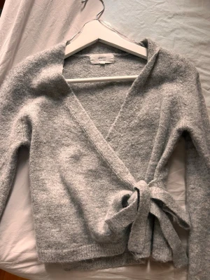 Grå omlottkofta från Gina Tricot - Mysig grå omlottkofta från Gina Tricot i ullblandning. Koftan har långa ärmar, knytskärp i midjan och en mjuk, fluffig känsla. Perfekt för lager på lager och ger en avslappnad men ändå stilren look.